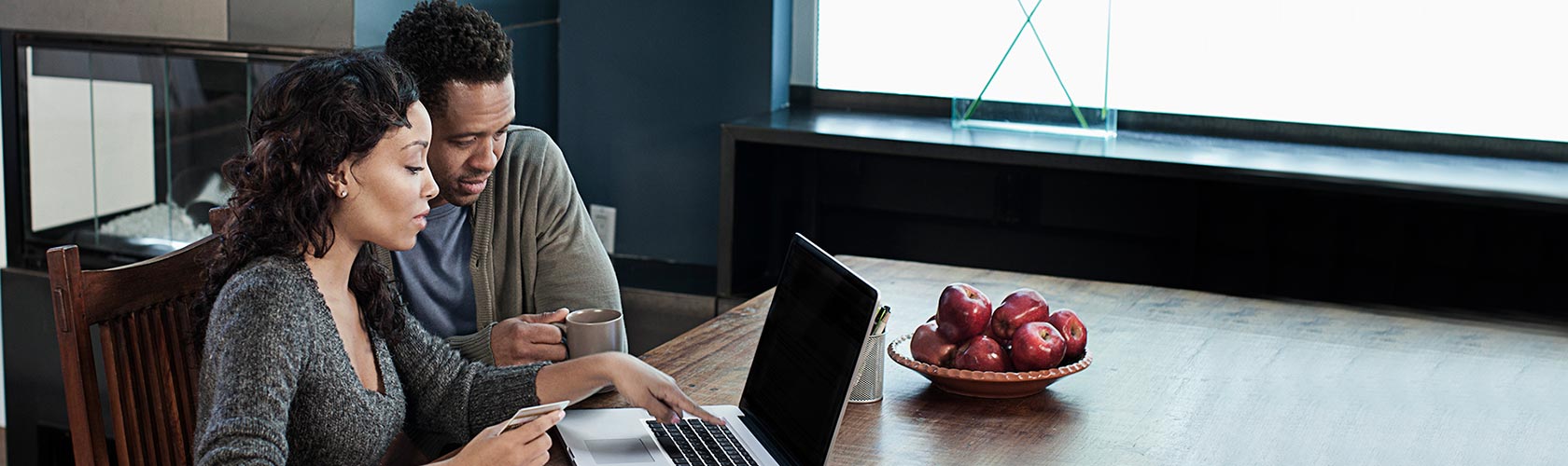 a husband and wife sit together at desk reviewing finances on laptop computer