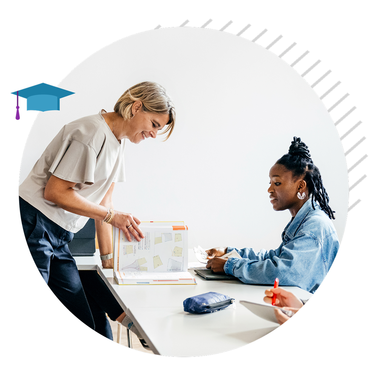 A teacher stands leafing through a book in front of a student sitting at a desk.
