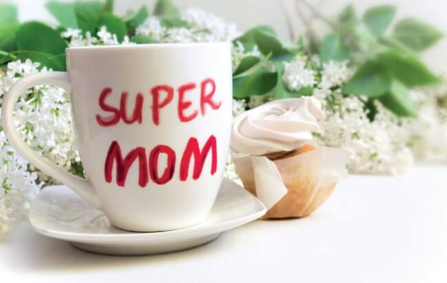 A coffee mug with text "Super Mom" and a cupcake sits on a table of flowers
