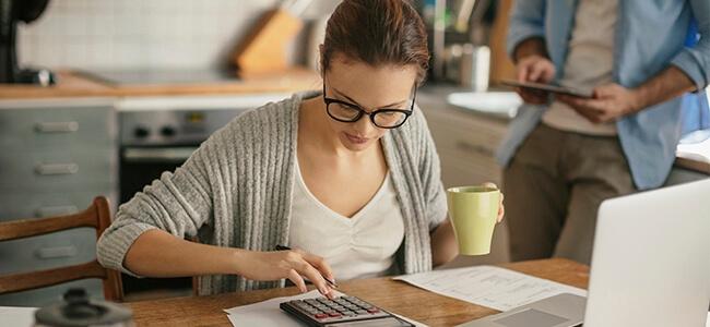 A woman sits at kitchen table with coffee, reviewing finances, working on calculator