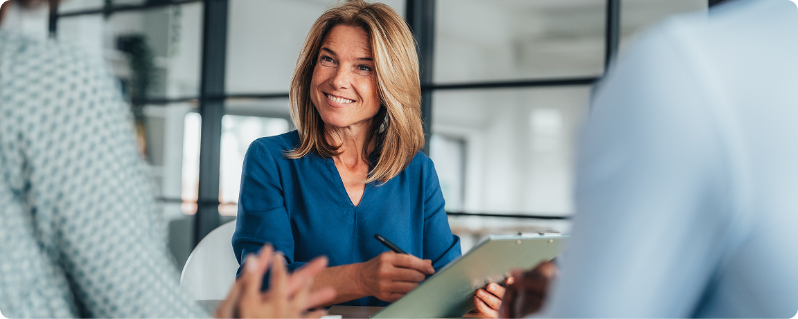 A woman smiles as she holds a clipboard and speaks to 2 individuals.