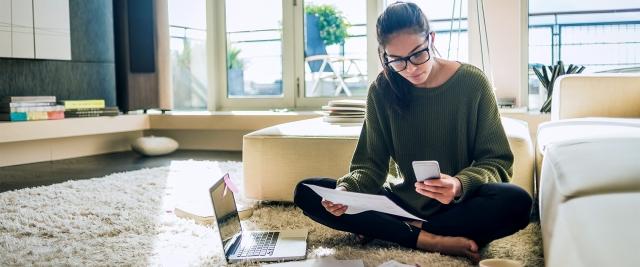 a woman sits on the floor of her living room with laptop reviewing paper bills