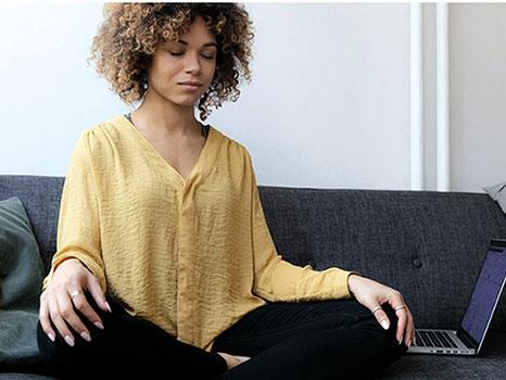 a woman sits on her couch in meditation pose with laptop nearby